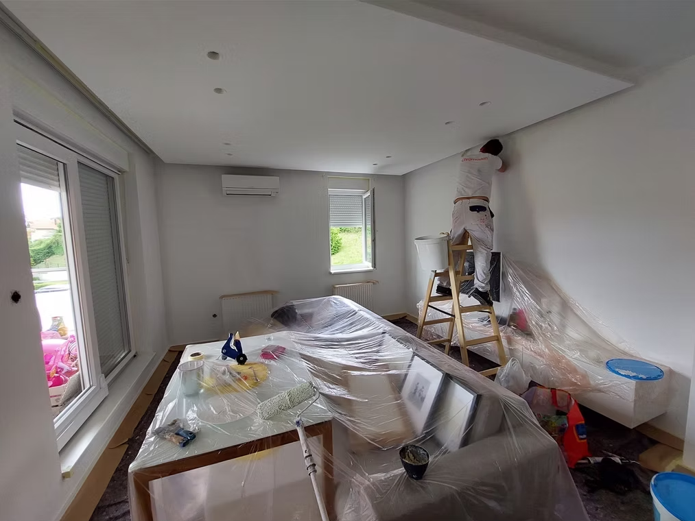 Slika pokazuje majstora koji kreči. U centru sobe nalazi se uredno poslagan namještaj zaštićen plastičnom folijom kako bi ga se sačuvalo od prljanja.